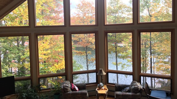 A room with large windows offering a view of autumn foliage and a lake. There are two chairs and a small table with a lamp. - big house on the private lake ,10 minutes to downtown and Lake Michigan (Traverse City)