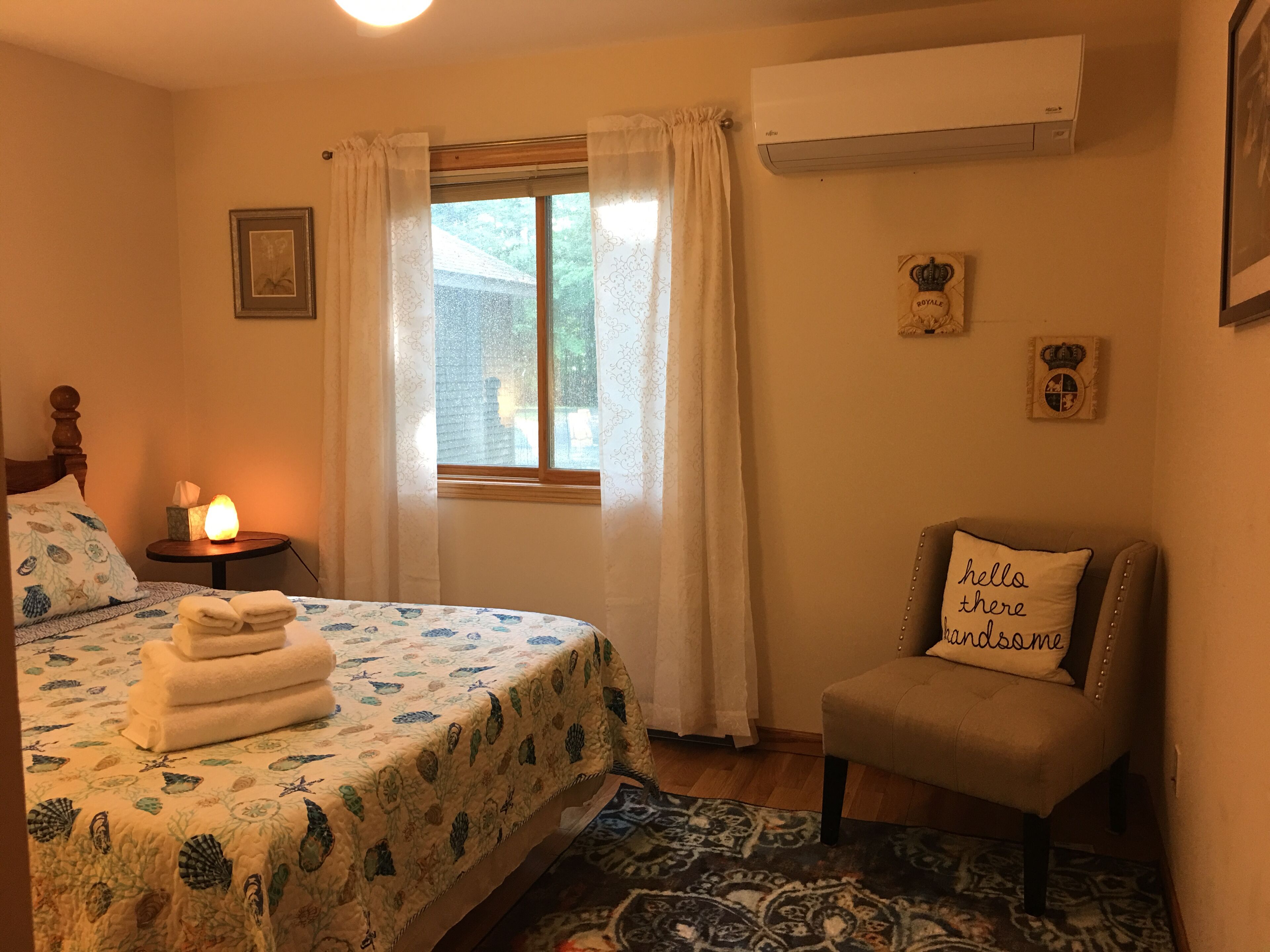 A bedroom with a bed, a bedside table with a lamp, a chair with a cushion, and a window with curtains.