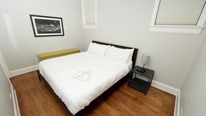 A neatly made bed with white bedding, a small bedside table with a lamp, and a framed picture on the wall. - Wrigley Field MVP Suite 1 Bedroom Apartment (Chicago)