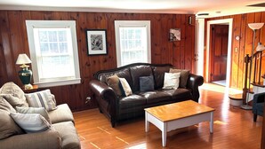 Ett vardagsrum med en soffa i läder, ett soffbord i trä och en beige divansoffa. Det finns två fönster med persienner, träpanel på väggarna och en dörr som leder till ett annat rum.