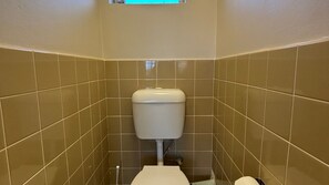 A small, enclosed bathroom with a toilet, a visible pipe, and a window above the toilet.