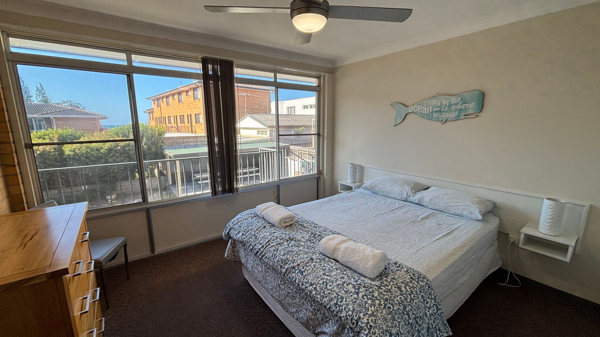 A bedroom with a large window, a ceiling fan, a bed with a patterned bedspread, and a wall-mounted fish decoration.