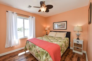 A bedroom with a bed, two bedside tables, a ceiling fan, a window with curtains, and a framed picture on the wall. - AMAZING VIEW! Fun, Pets Welcome, Steps from Beach (St George Island)