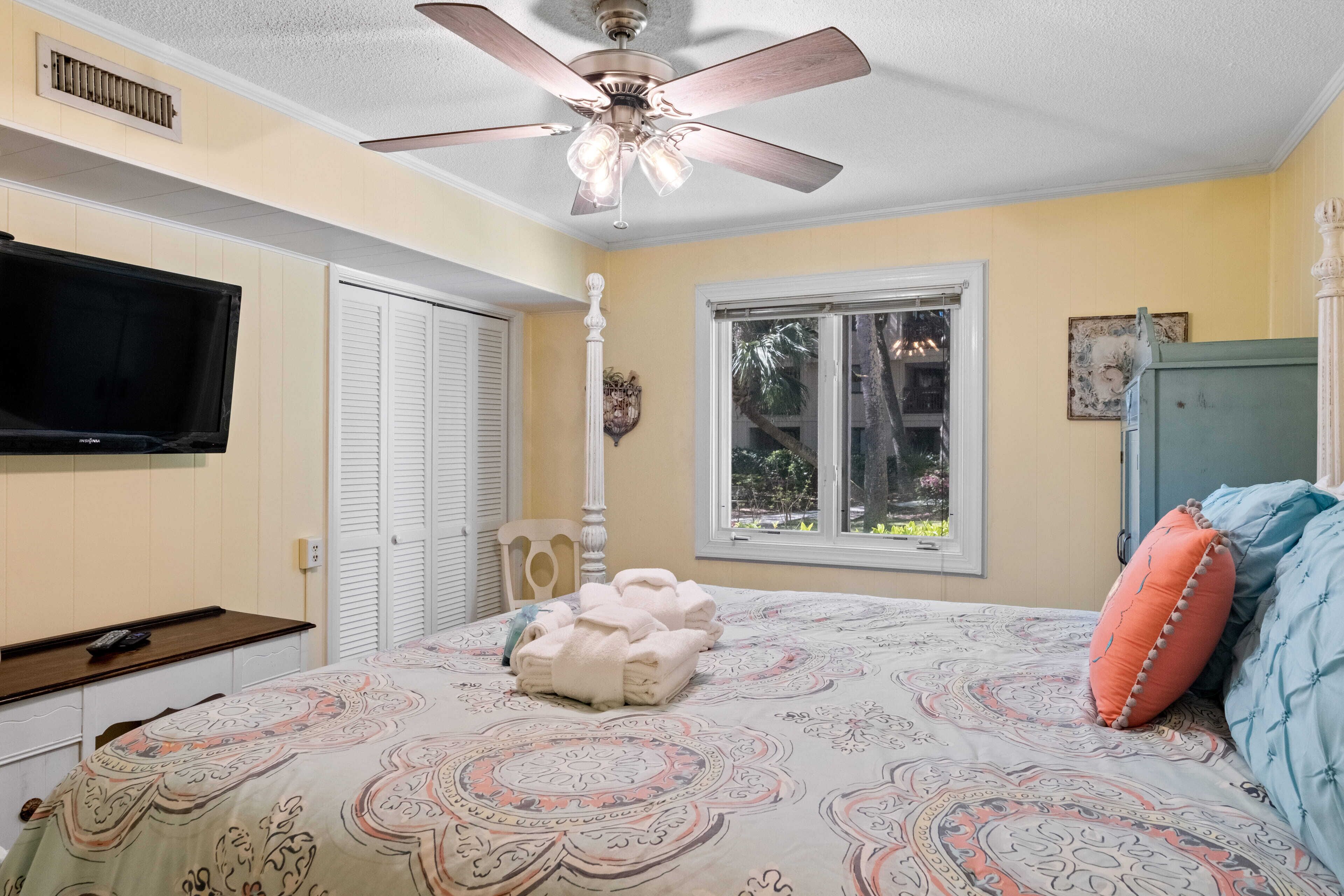 A bedroom with a bed, a ceiling fan, a TV, a window, and a door.
