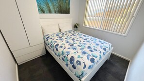 A small, single-bed room with a floral bedspread, a white headboard, a window with blinds, and a painting on the wall. - Chriscliffs Hat Head (HAT HEAD)
