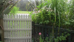 Una cerca blanca de piquetes con un portón de metal, rodeada de exuberante follaje y plantas florecientes.
