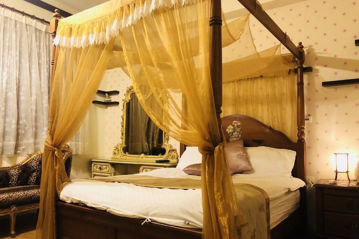 A four-poster canopy bed with a wooden frame and gold drapes, a nightstand with a lamp, and a leopard print chair.