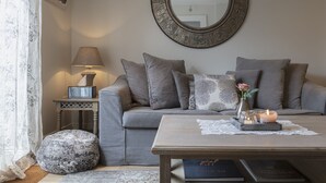 A cozy living room with a grey sofa, a round mirror, a small table with candles, and a patterned ottoman. - Shrubbery Mews 5 * Apt 2 (Kenmare)