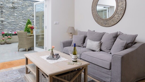 A living room with a grey sofa, a coffee table with a bottle and glass, a wicker chair, and a stone wall outside. - Shrubbery Mews 5 * Apt 2 (Kenmare)