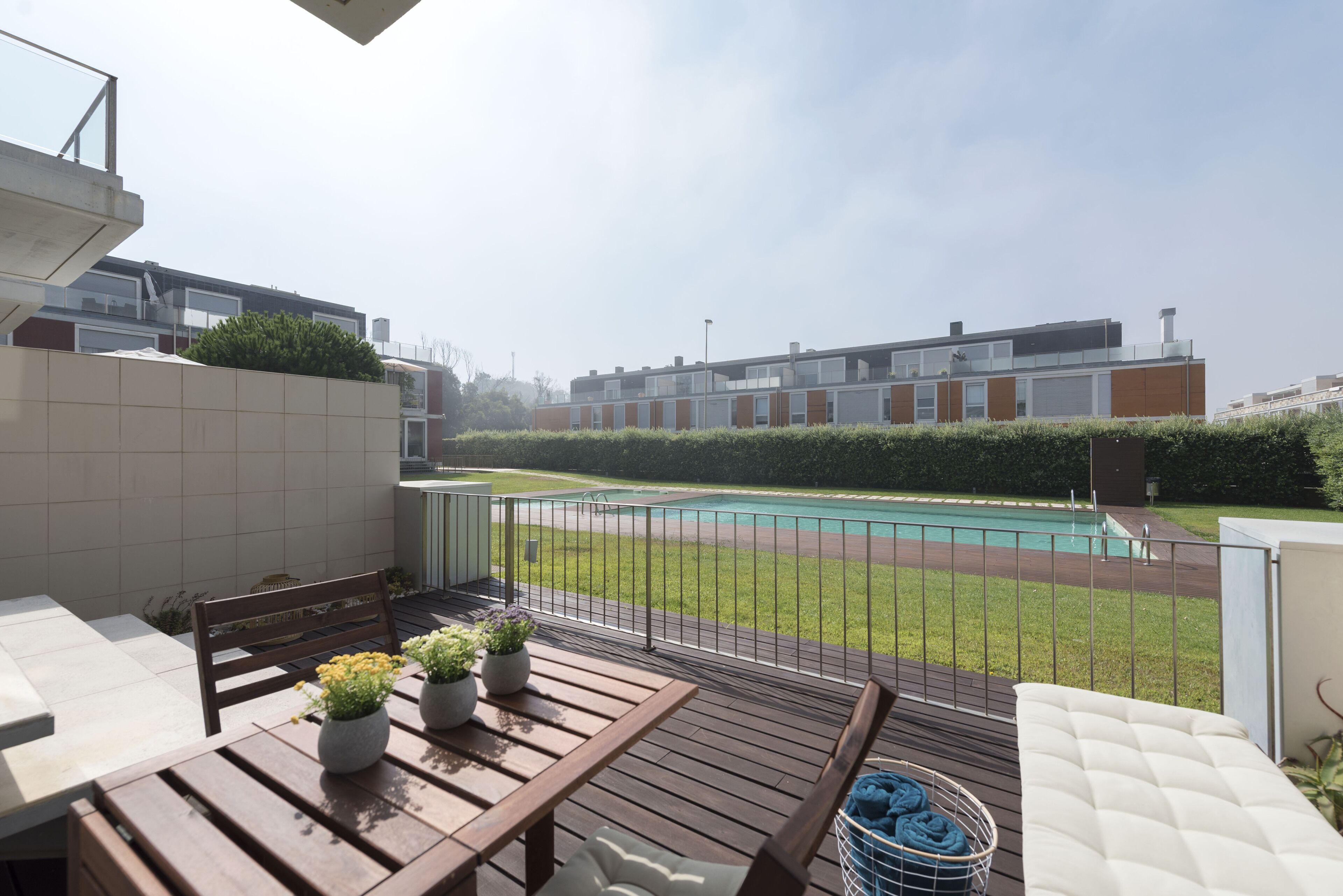 Un balcón con mesa de madera, plantas en macetas y vistas a una piscina y edificios residenciales.