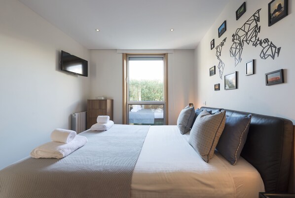 A modern hotel room with a large bed, a flat-screen TV, a world map artwork, and a view of greenery through the window.