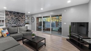 A modern living room with a grey sofa, a coffee table, a TV on a stand, and a stone wall. - Lester Place: Retro-Inspired Artist’s Escape with Pool! (Tucson)