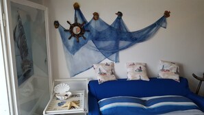A bedroom with a bed, pillows with lighthouse prints, a wall hanging with a ship's wheel, and a small white table with a shell and starfish. - THE FISHERMAN'S HOUSE (Ospedaletti)