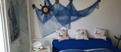 A bedroom with a bed, pillows with lighthouse prints, a wall hanging with a ship's wheel, and a small white table with a shell and starfish.