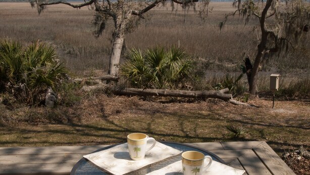 A round glass table with two white mugs on white napkins, set on a wooden deck with a view of a grassy field and trees.