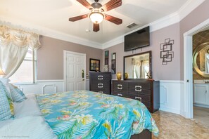 Une chambre à coucher avec un lit, un ventilateur de plafond, une commode, un téléviseur et une porte menant à la salle de bain.