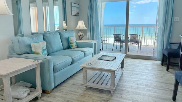 A living room with a sofa, coffee table, and chairs, a balcony with a view of the beach.