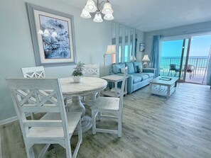 A living room with a round dining table, white chairs, a sofa, and a balcony with a view of the ocean. - Fresh Updates | Harbor House B6 - Awesome Beach Front Views, Free WiFi, Prime location (Gulf Shores)