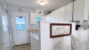 A kitchen with white cabinetry, a clock, a framed sign reading 'Seaside Villas,' and a window with blinds.