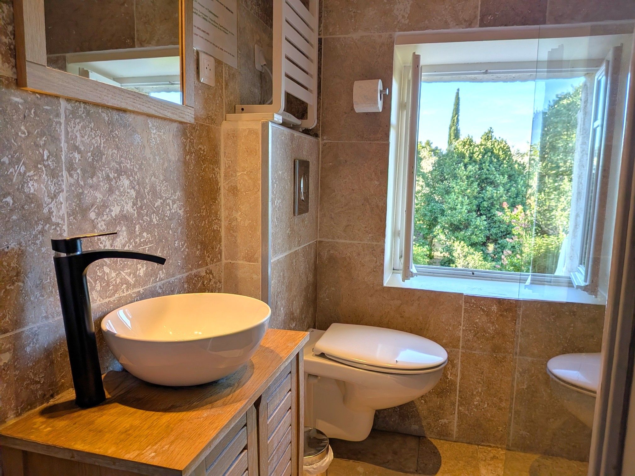 A bathroom with a large white sink, a toilet, and a window with a view of greenery.