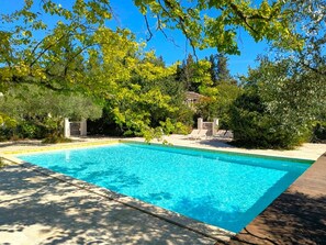 A rectangular swimming pool with a tiled deck, surrounded by lush greenery and trees.