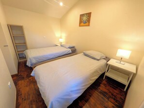 A small, well-lit room with two beds, a nightstand with a lamp, and a wooden floor.