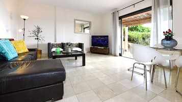 Un salon moderne avec un canapé en cuir noir, une table basse, un téléviseur et un coin repas avec des chaises blanches.