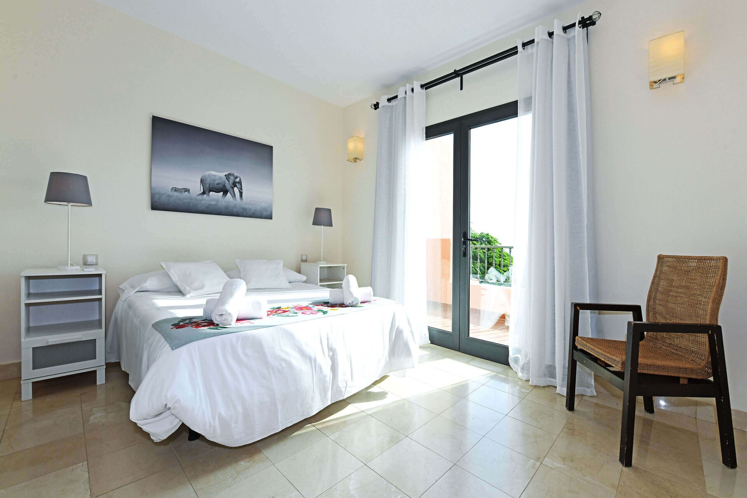 Une chambre d’hôtel avec un lit, deux lampes de chevet, une chaise, un tableau représentant un éléphant et une porte-fenêtre coulissante donnant sur un balcon.