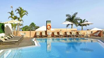 A swimming pool with lounge chairs and umbrellas, surrounded by palm trees and a clear sky.
