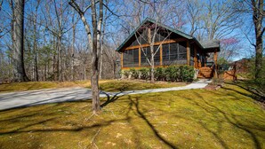 A log cabin with a large window, surrounded by trees and a grassy yard.