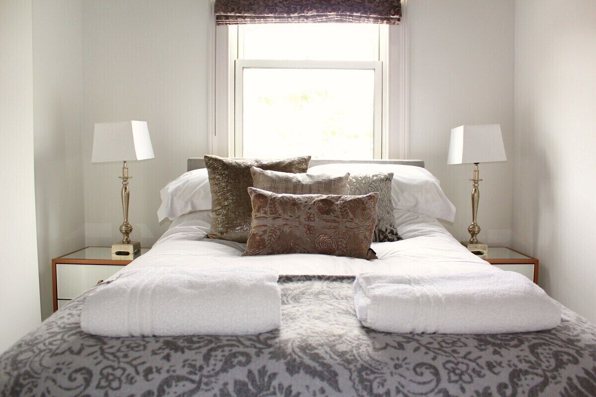 A neatly made bed with white linens and patterned pillows, two bedside lamps, and a window with a view.