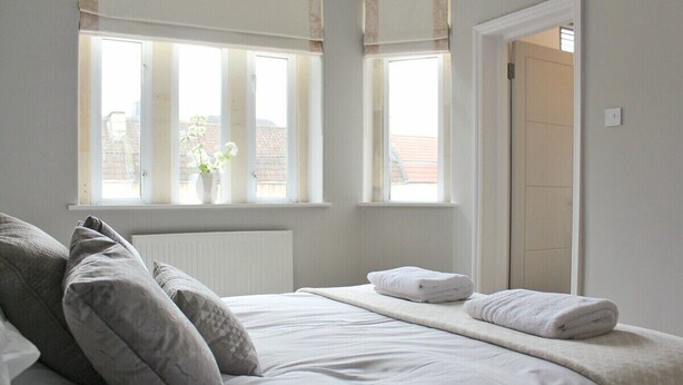A bedroom with a bed, pillows, a radiator, and two windows. - Lovely Central Bath House- w/ Parking (CG) (Bath)