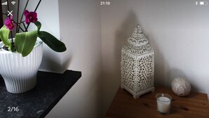 A white ceramic pot with a plant on a dark countertop, a white intricately designed lantern, and a candle on a wooden table against a plain wall.
