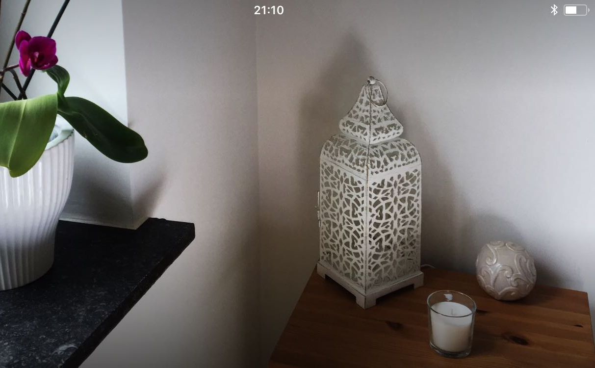 Ein weißer Keramiktopf mit Pflanze auf einer dunklen Arbeitsplatte, ein kunstvoll gestalteter, weißer Laternenleuchter und eine Kerze auf einem Holztisch vor einer schlichten Wand.