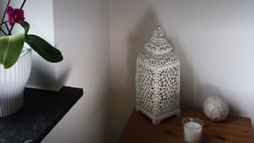 Ein weißer Keramiktopf mit Pflanze auf einer dunklen Arbeitsplatte, ein kunstvoll gestalteter, weißer Laternenleuchter und eine Kerze auf einem Holztisch vor einer schlichten Wand.