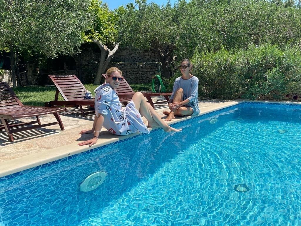 Une piscine où deux personnes sont assises au bord, entourées de chaises longues et de verdure.