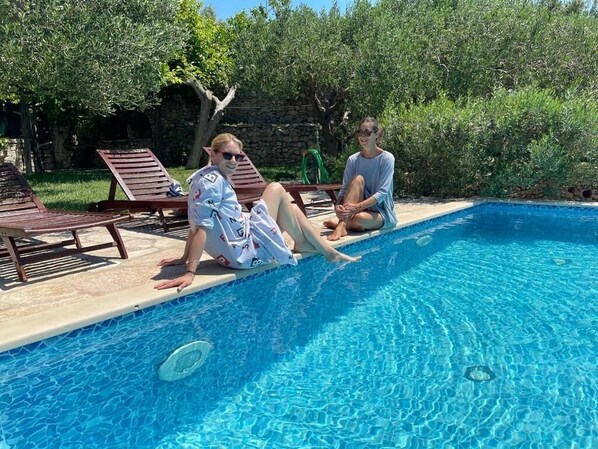 A swimming pool with two people sitting on the edge, surrounded by lounge chairs and greenery. - Holiday Home in Bol with Seaview, Terrace, Air condition, WIFI (120-1) (Bol)