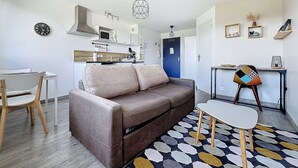 Un salon moderne avec un canapé marron, une petite table blanche et un tapis coloré. On aperçoit, au fond, une cuisine avec des meubles blancs et une porte bleue.