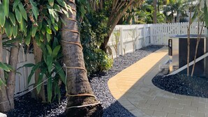 A pathway lined with palm trees and a white fence, leading to a building with a brick exterior. - 1 Bedroom Suite in a duplex house- private heated pool for you- walk to beach  (Lauderdale-By-The-Sea)