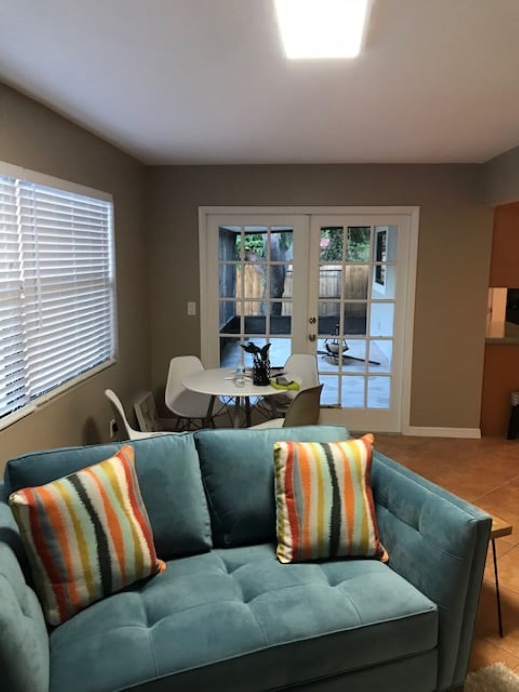 A living room with a teal sofa, striped pillows, a dining table, and a view through a glass door to an outdoor area.