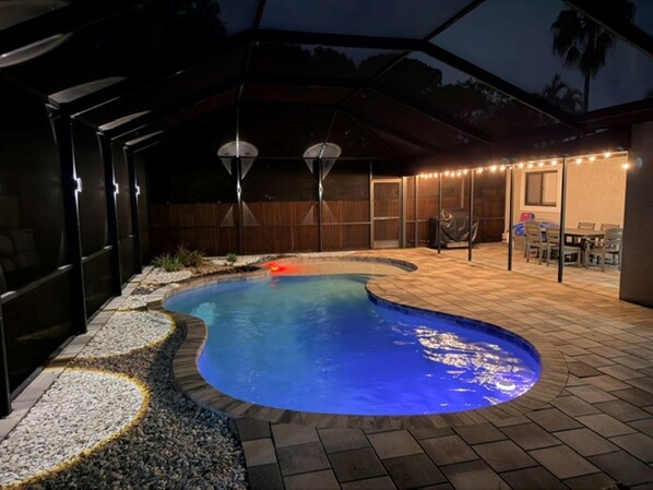 An outdoor swimming pool with a curved edge, surrounded by a paved deck and a covered seating area with string lights.