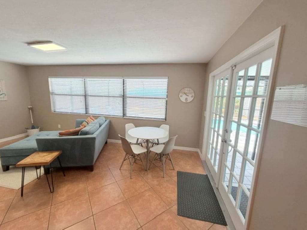 A living room with a sectional sofa, a small table, and a dining area with chairs. There is a clock on the wall and a window with blinds.