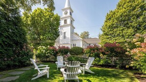 A white church with a steeple and clock, surrounded by lush greenery and flowering bushes.