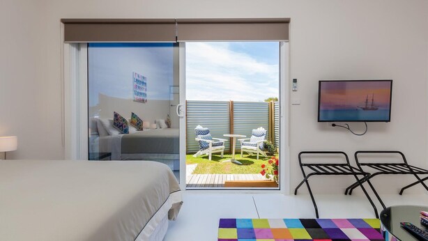 A hotel room with a bed, a TV, a balcony with outdoor furniture, and a painting on the wall.
