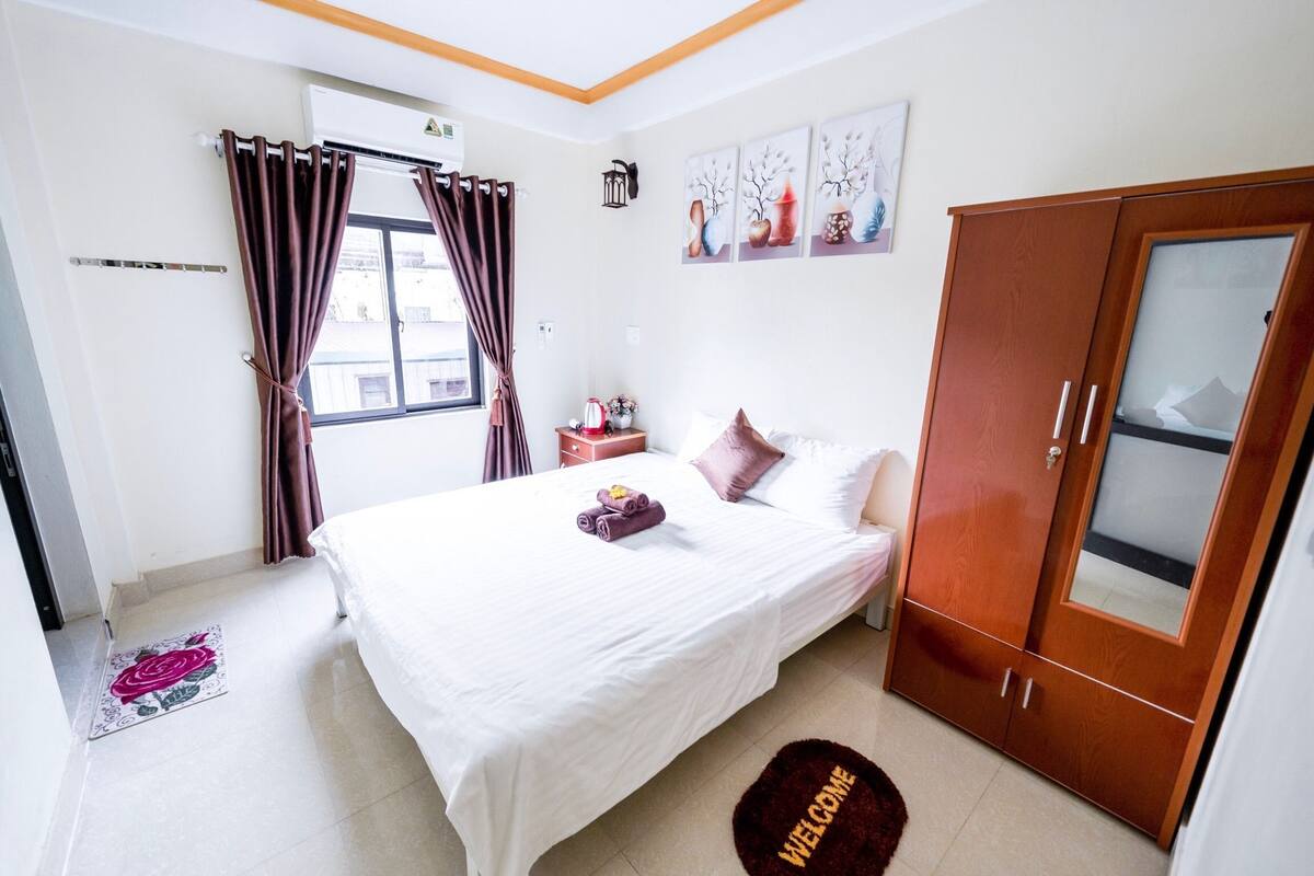 A hotel room with a bed, a wardrobe, a nightstand, a window with curtains, and artwork on the wall.