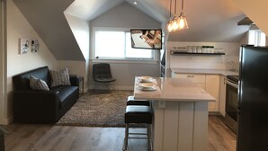Una cocina moderna con una isla central, un comedor con sofá negro y una silla, y un amplio ventanal que proporciona luz natural.