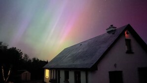 Una casa de techo inclinado y chimenea, bajo un cielo nocturno con aurora boreal visible.