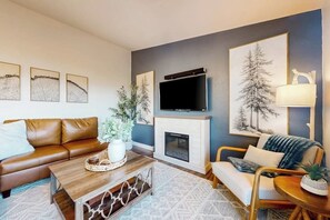 A living room with a brown leather sofa, a wooden coffee table, a fireplace, a flat-screen TV, and framed artwork on the walls. - Spacious rental with furnished deck & great views of the lake (Garden City)
