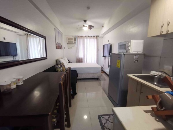 A compact hotel room with a bed, a small kitchenette, a dining table, and a ceiling fan.