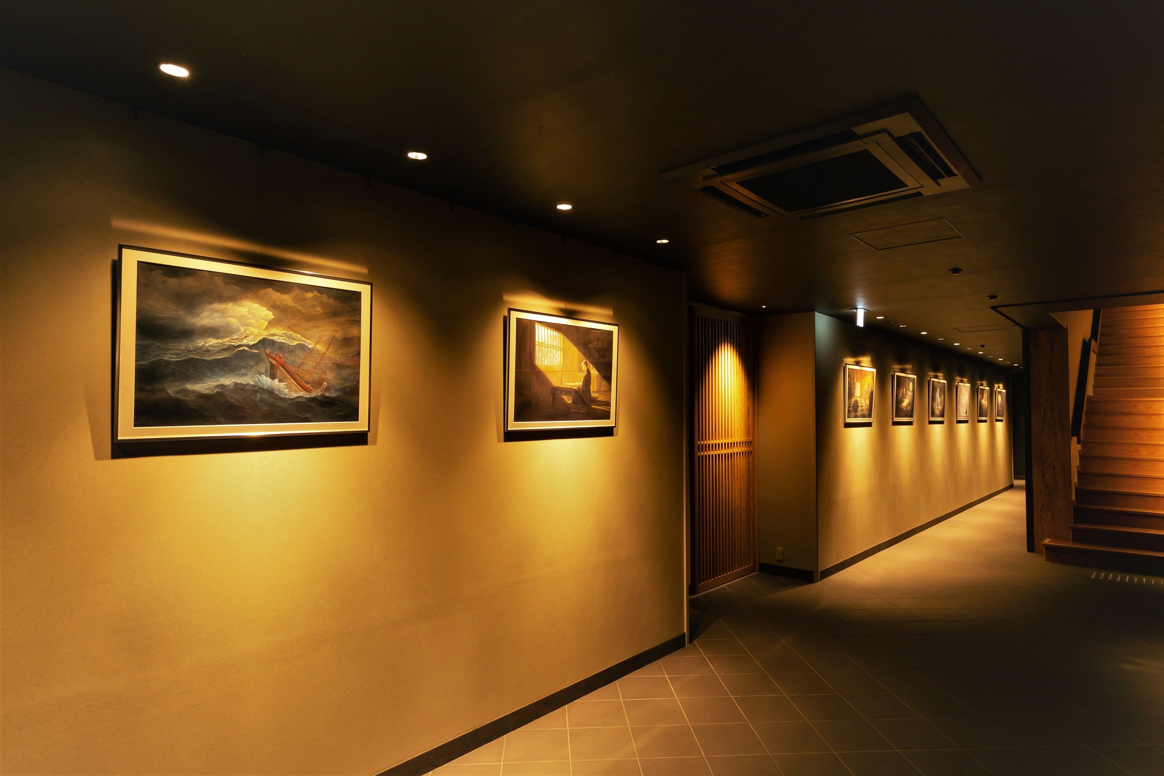 A hallway with framed artwork on the walls, a wooden staircase, and recessed lighting.